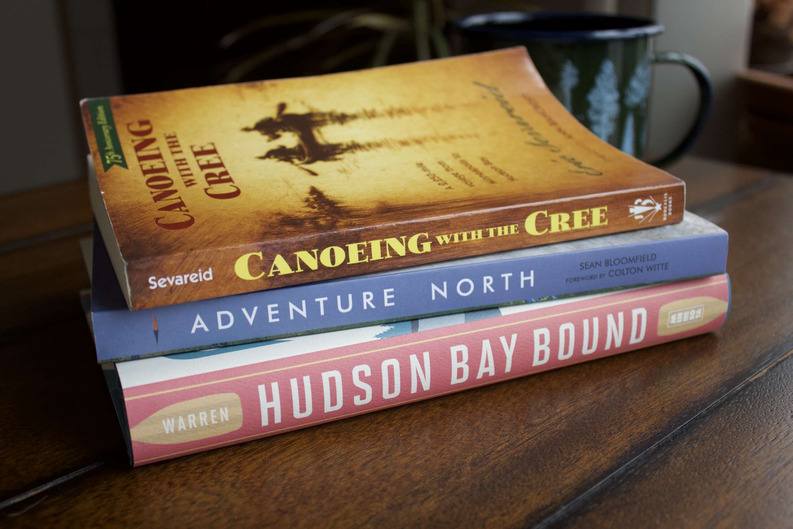 Three books, "Canoeing with the Cree", "Adventure North", and "Hudson Bay Bound" sit in a stack on a dark colored table